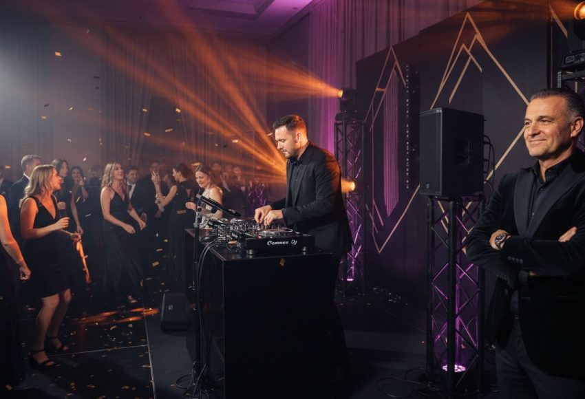 DJ, der an einem Mischpult auf einer Veranstaltung spielt, während das Publikum zuschaut und feiert.