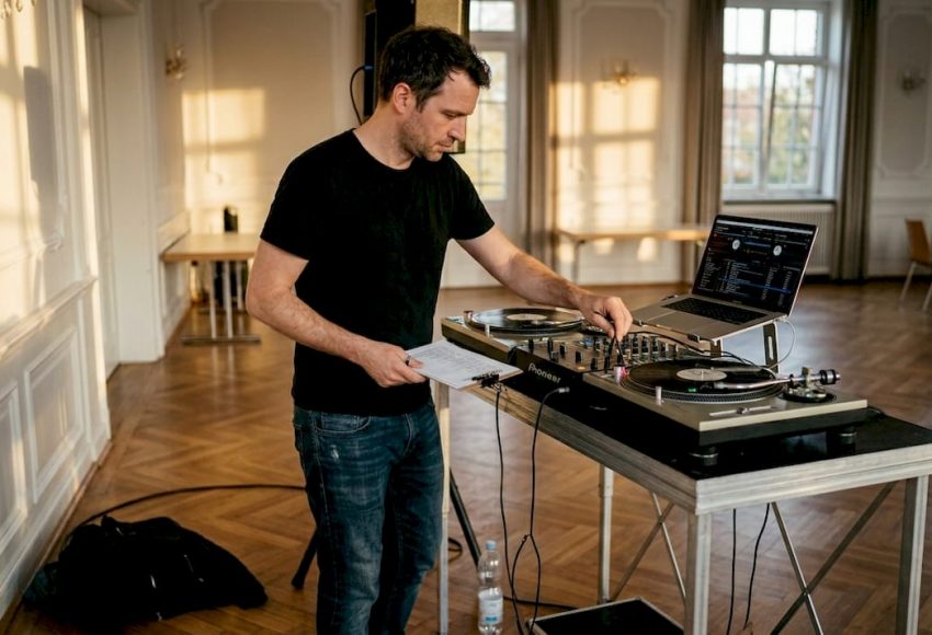 Warum Spezialisierung auf München bei Events entscheidend ist 10 Ein Mann in schwarzem T-Shirt arbeitet an einem DJ-Setup mit Plattenspielern und Laptop, leiser Sonnenlichtstrahl fällt auf einen Holzfußboden in einem großen Saal in München