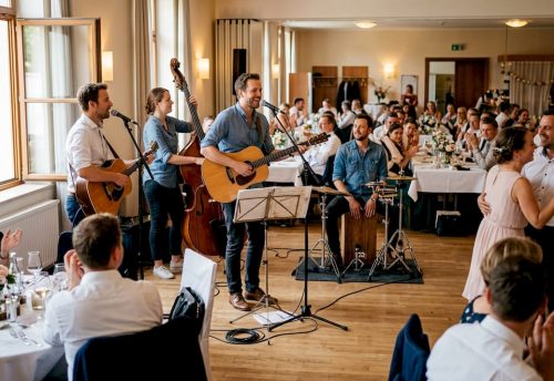Musikauswahl für Events: Emotionale Verbindung & Stimmung 3 Eine Live-Band spielt auf einer Hochzeit, der Sänger mit Akustikgitarre und weitere Musiker sorgen für eine emotionale Atmosphäre, präsentiert von Top DJ München