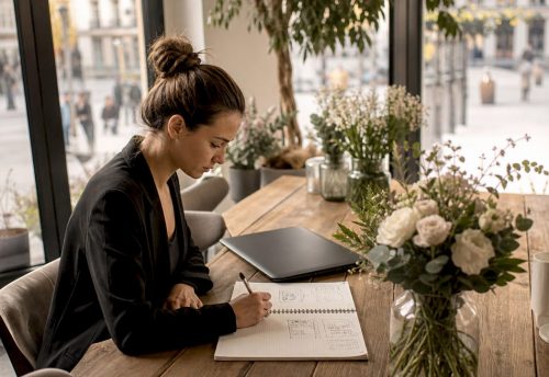 Was bedeutet maßgeschneidertes Event? Alles Wichtige 5 Eine Eventplanerin entwickelt am Holztisch ein maßgeschneidertes Eventkonzept und notiert Ideen neben Blumen, Laptop und Stadtblick im hellen Raum.