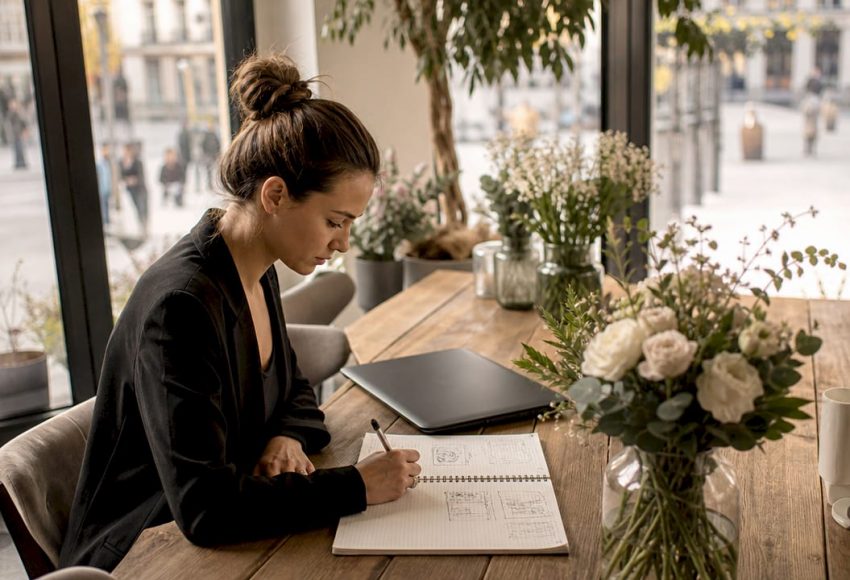 Was bedeutet maßgeschneidertes Event? Alles Wichtige 1 Eine Eventplanerin entwickelt am Holztisch ein maßgeschneidertes Eventkonzept und notiert Ideen neben Blumen, Laptop und Stadtblick im hellen Raum.