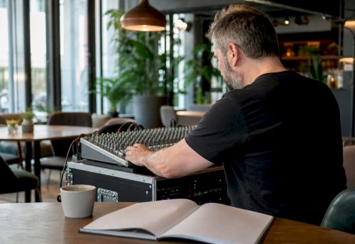 Hochwertige Soundanlage mieten: Guide für München 4 Ein Tontechniker justiert an einem Mischpult in einem Café die Anlage für ein Event, während vor ihm ein Notizbuch und eine Tasse auf dem Tisch stehen.