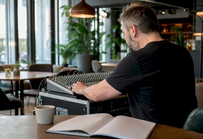 Hochwertige Soundanlage mieten: Guide für München 4 Ein Tontechniker justiert an einem Mischpult in einem Café die Anlage für ein Event, während vor ihm ein Notizbuch und eine Tasse auf dem Tisch stehen.