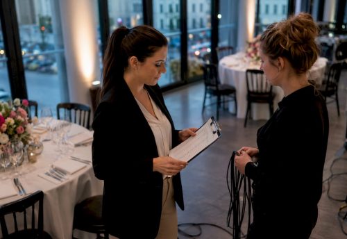Event-Entertainment-Trends 2025: Der Münchner Praxisguide 6 Eine Eventplanerin spricht in einem festlich gedeckten Saal mit einer Kollegin und prüft eine Checkliste für den Aufbau in München.