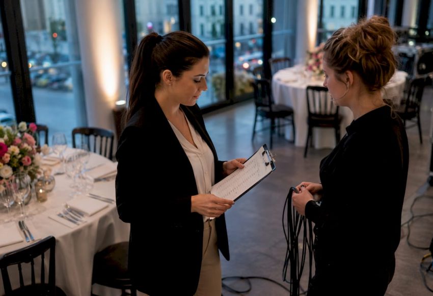Event-Entertainment-Trends 2025: Der Münchner Praxisguide 6 Eine Eventplanerin spricht in einem festlich gedeckten Saal mit einer Kollegin und prüft eine Checkliste für den Aufbau in München.
