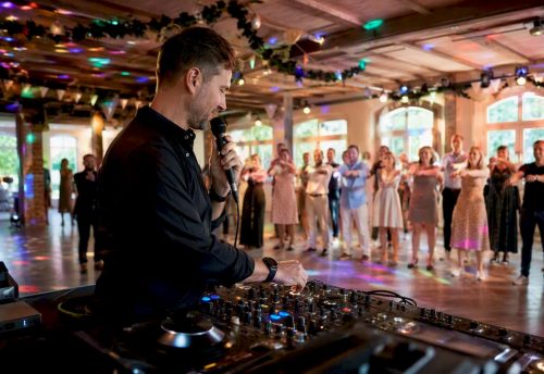 Die besten Partyspiele 2025 für Events in München 6 DJ Mike Lindström spricht ins Mikrofon und steuert ein Partyspiel vor einer Gruppe von Gästen in einer Eventlocation.