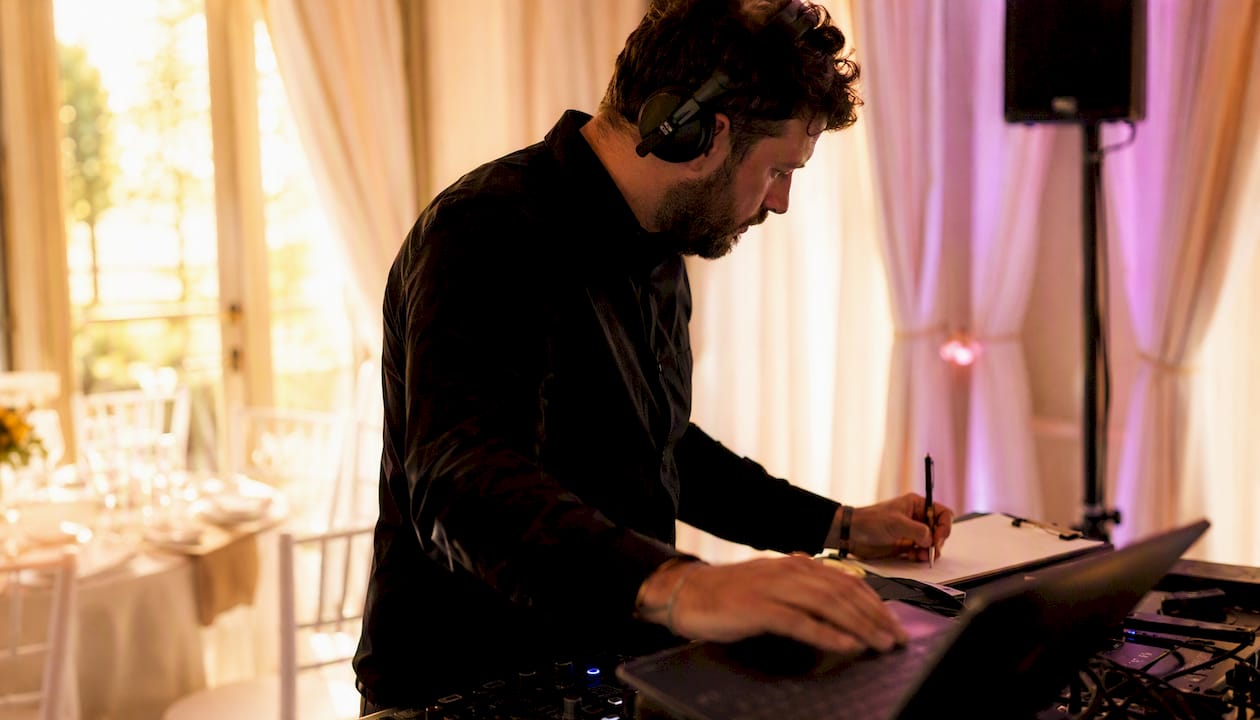 Ein Hochzeits-DJ mit Kopfhörern notiert Songs am Mischpult und steuert den Laptop bei einer Feierlocation.